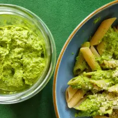 Pesto z liści rzodkiewki w słoiku obok talerza penne z tym zielonym sosem i startym parmezanem.