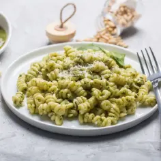 Pyszny makaron z pesto ze słoika, posypany parmezanem. Obok kawałki sera i garść orzeszków piniowych.