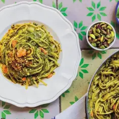 Pyszne rodzaje makaronu z pesto pistacjowym i kawałkami łososia, posypane orzechami. Obok miseczka pistacji i listki bazylii.