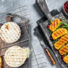 Dwa okrągłe sery na grilla, jeden lekko roztopiony, obok grzanki i grillowane kawałki dyni z granatem.