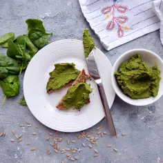 Pesto ze szpinaku na grzankach, obok miseczka z pastą i świeże liście szpinaku.