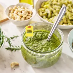 Pesto z rukoli w słoiku, obok miseczki z rukolą, orzechami nerkowca i makaronem. Idealne do letnich dań.
