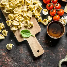 Surowe ravioli i tortellini na desce, obok pomidorków i miseczki z sosem.