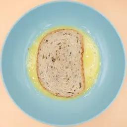 Kromka chleba z ziarnami zanurzona w roztrzepanym jajku na niebieskim talerzu. Przygotowanie do pysznych jajochlebków.