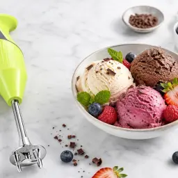 Zielony blender ręczny obok miski z lodami waniliowymi, czekoladowymi i jagodowymi, ozdobionej owocami. Idealne do najprostszych lodów domowych.