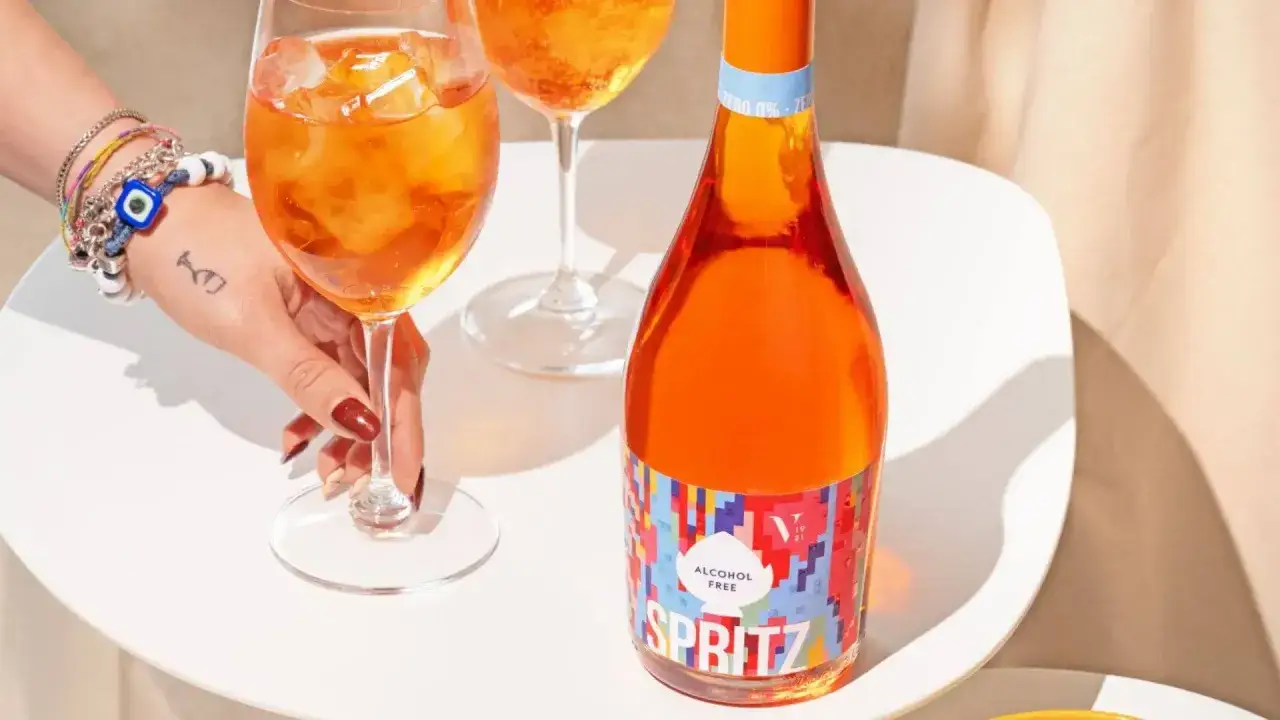 Orzeźwiający aperol bezalkoholowy w szklance z lodem, obok butelki z kolorową etykietą. Idealny na letnie popołudnie.