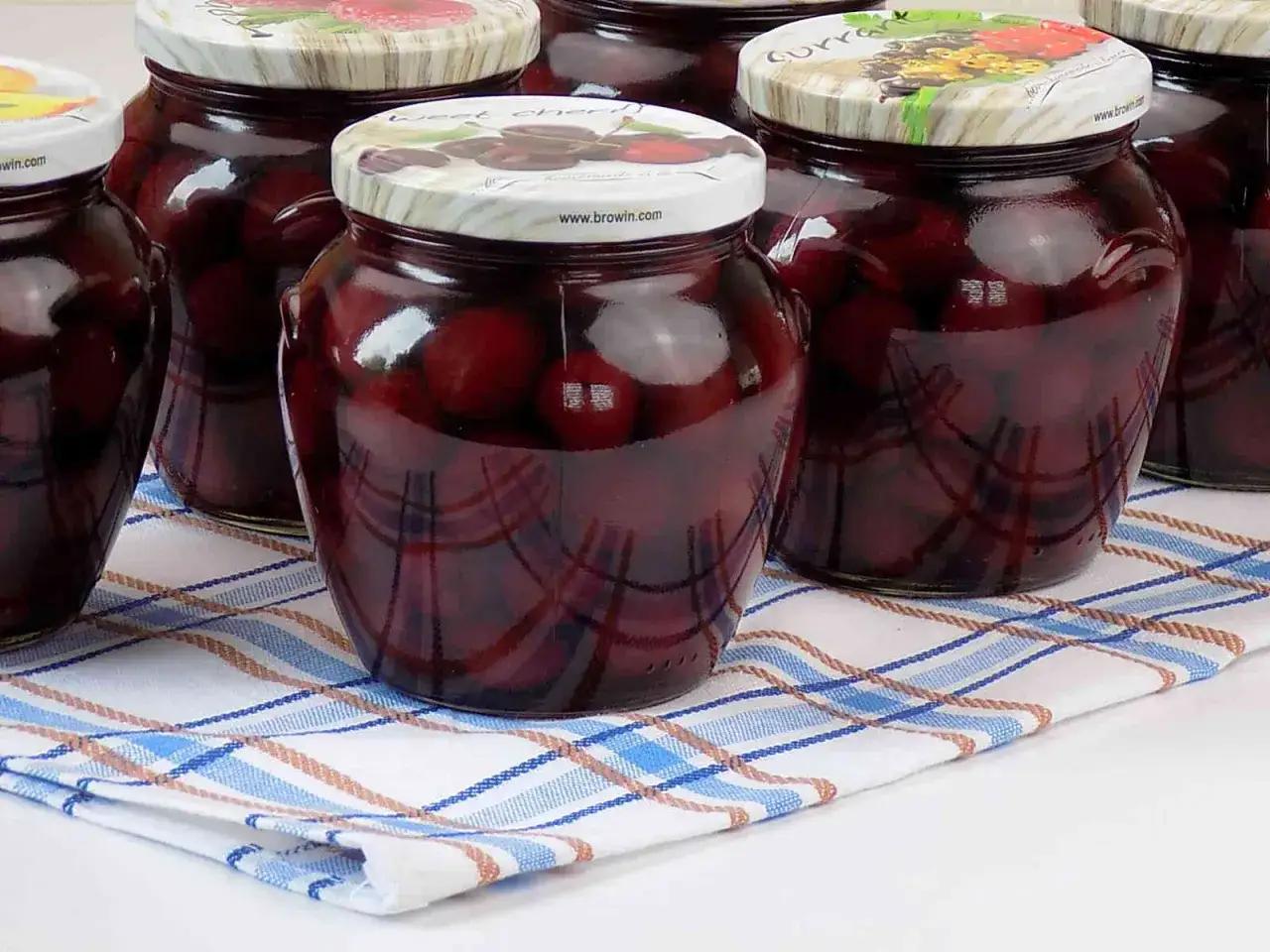 Słoiki z kompotem z czereśni do słoików, gotowe na zimę. Ciemne owoce w słodkim syropie, pięknie prezentują się na kratkowanej serwetce.