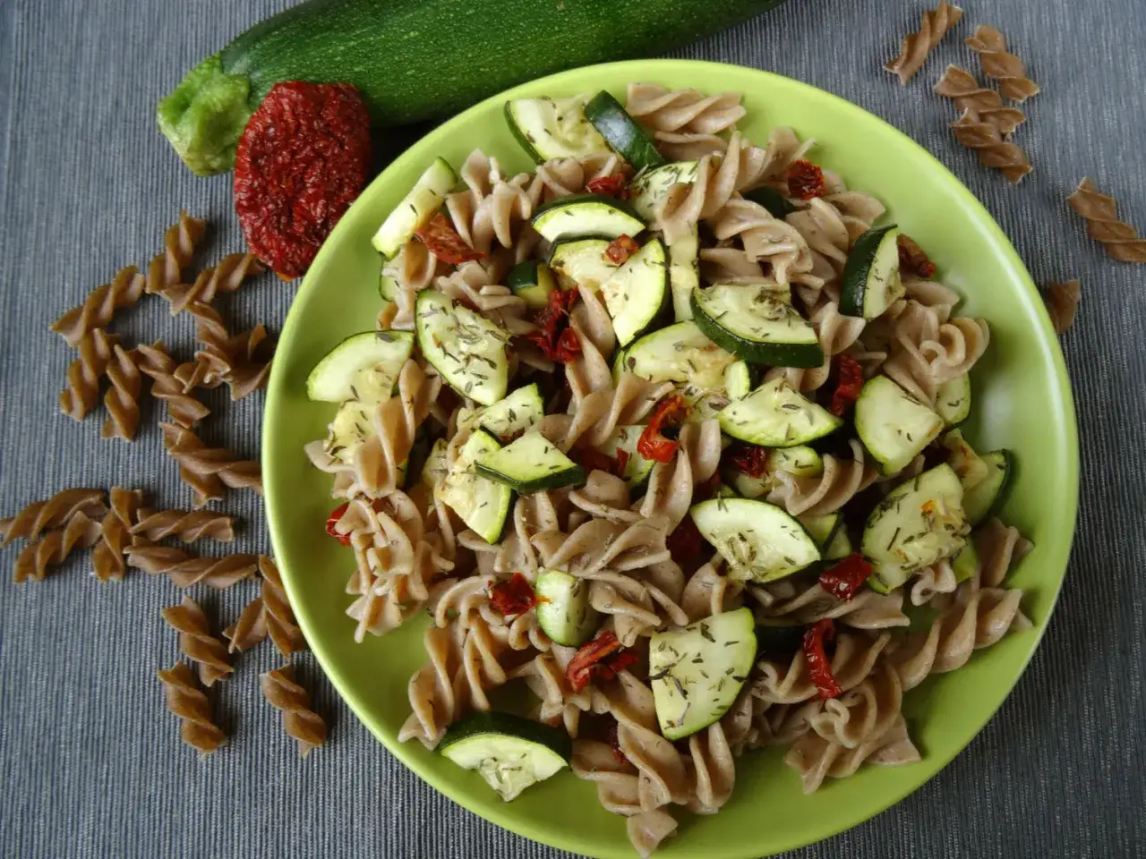 Makaron z cukinią i pomidorami, posypany ziołami, na zielonym talerzu. Obok leżą surowe składniki i makaron fusilli.