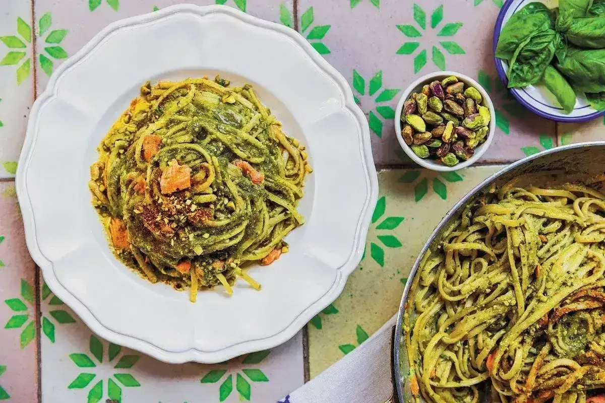 Pyszny makaron z pesto ze słoika, posypany pistacjami i kawałkami suszonych pomidorów. Obok miseczka z pistacjami i listkami bazylii.