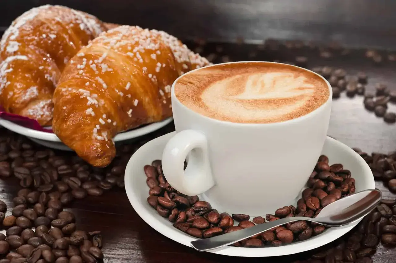Idealne włoskie śniadanie: cappuccino z pianką i dwa rogaliki posypane cukrem, otoczone ziarnami kawy.
