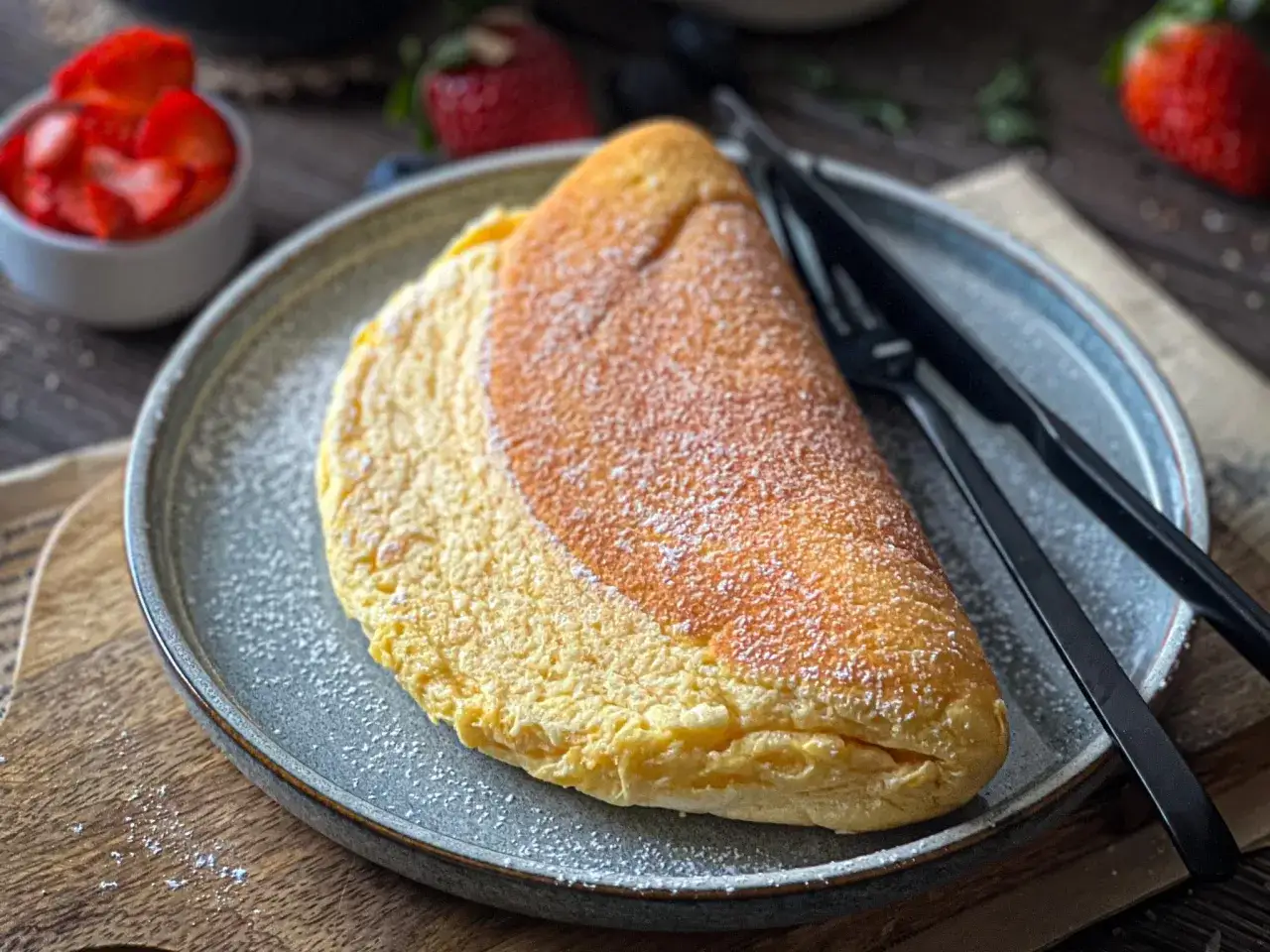 Puszysty omlet na talerzu, posypany cukrem pudrem. Idealny przepis na omlet, który zachwyci każdego. Obok czarne sztućce i miseczka z truskawkami.