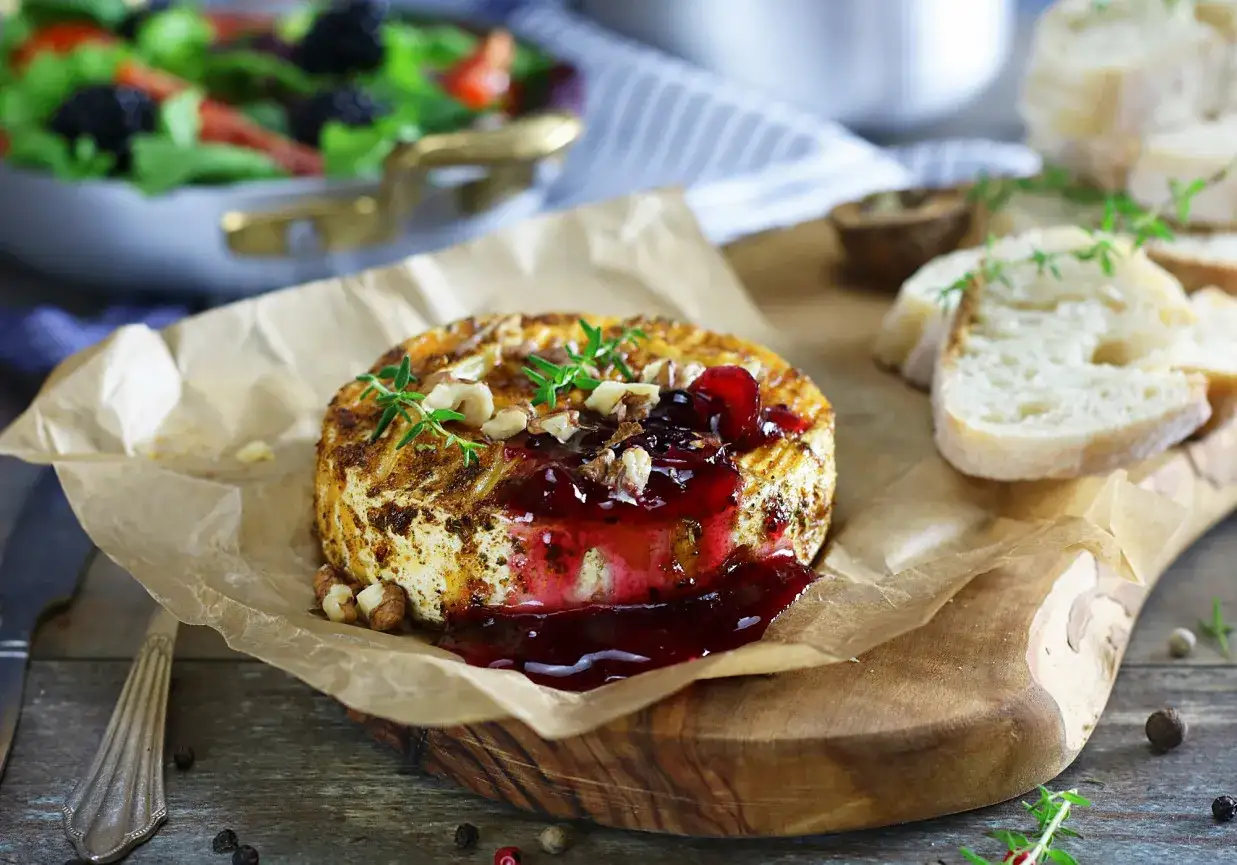 Pyszny camembert z grilla, polany konfiturą z owoców leśnych, posypany orzechami i gałązkami tymianku. Obok kromki chleba i sałatka.