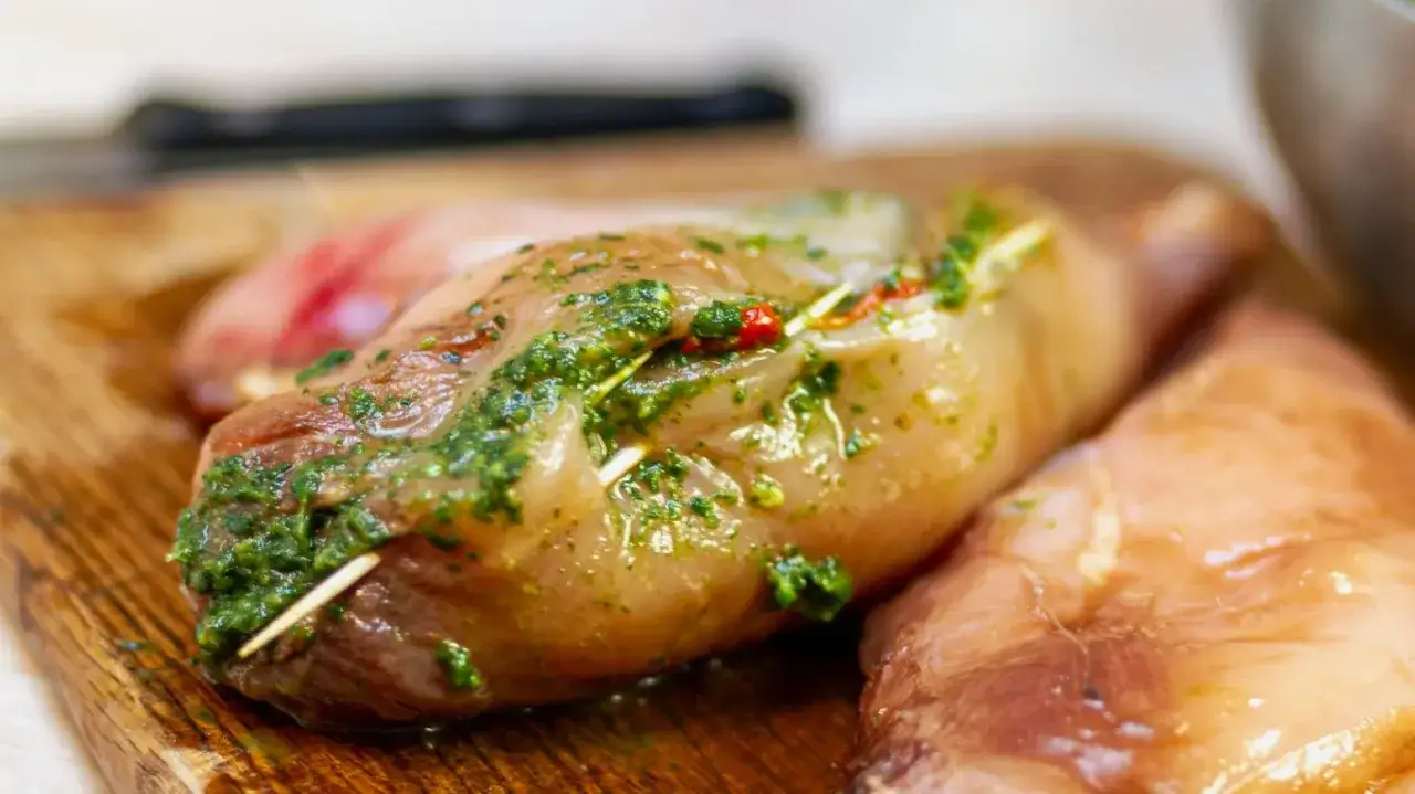 Surowe piersi kurczaka na desce, jedna nadziana zielonym pesto z pomidorkami.