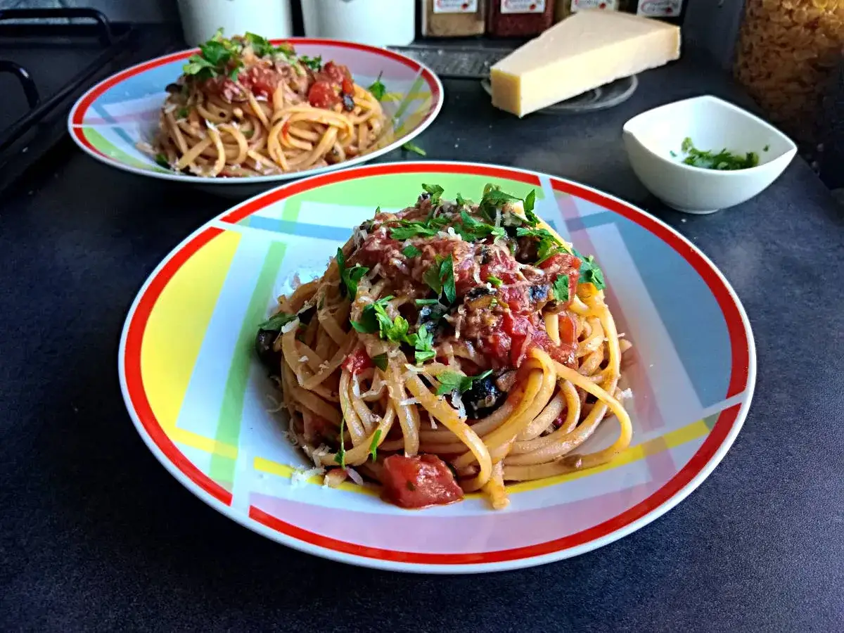 Dwa talerze spaghetti z pomidorowym sosem, oliwkami i parmezanem. Obok kawałek sera i miseczka z ziołami.