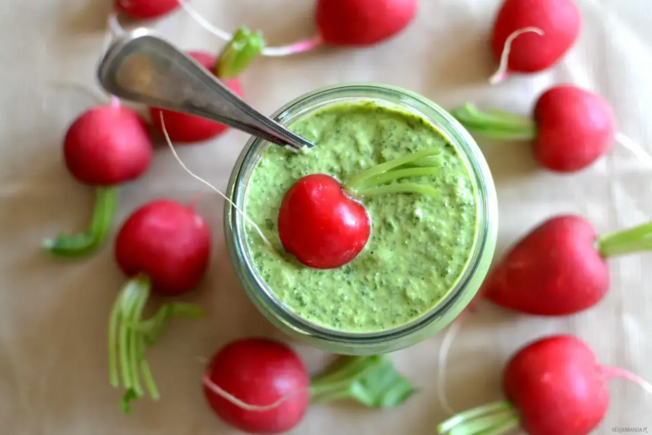 Słoik z zielonym pesto z liści rzodkiewki, ozdobiony jednym rzodkiewkowym serduszkiem. Wokół rozłożone są świeże rzodkiewki.