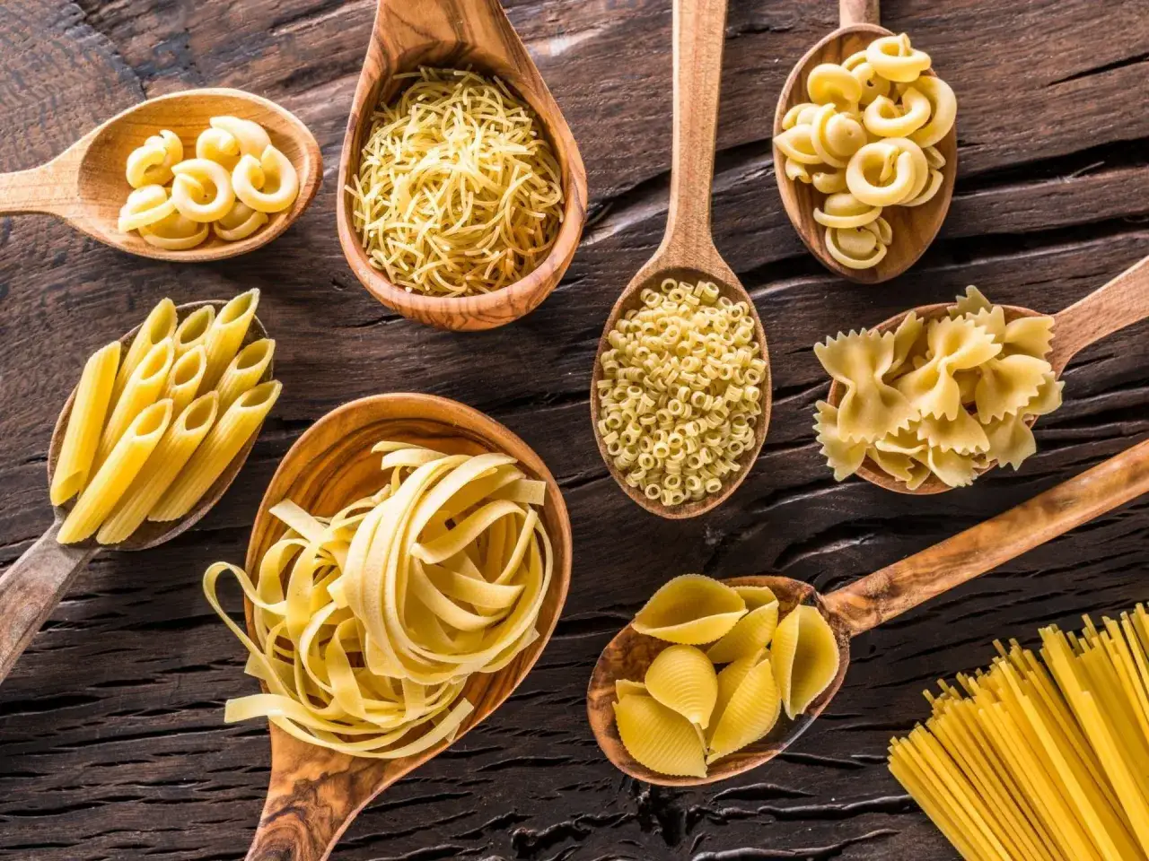 Drewniane łyżki pełne różnych rodzajów makaronu: penne, farfalle, spaghetti, conchiglie, anellini, vermicelli i inne.
